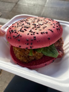 pink burger bun with black sesame seeds on top, filled with two large falafel. spinach leaf and small piece of red pepper poking out.