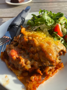 Square of lasagne with lentil and tomato mixture spilling out, and green salad with a cherry tomato quarter. knife and fork resting on side of plate.
